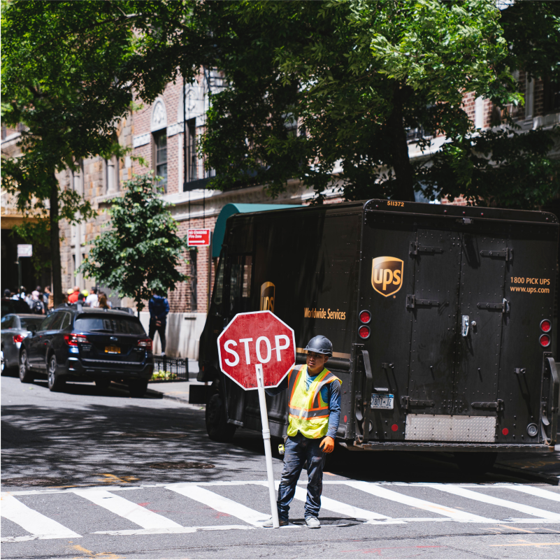 A man wearing a high vis jacket whilst holding a stop sign. Behind him is a parked UPS van.